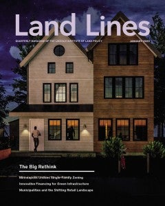 “Cyclist Centered Triplex” by Laura Stene, 2019. Credit: Courtesy of Designing for Minneapolis 2040 Studio, Dunwoody College of Technology, Minneapolis. A photograph of a modern multi-family, three-story home at night with two peaked roofs, many windows, and wood shingles. A man is entering the house and at least one person can be seen dimly inside the house. There are bushes and a bike rack, with a bike parked at it, in front of the home and trees and the night sky behind it.