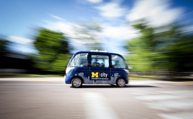 This image shows a street with a blue driverless shuttle in the middle. Blurred trees sit in the background of the image.