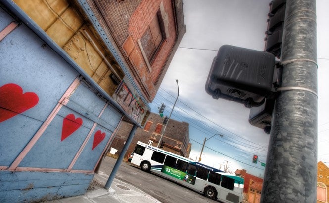 Photograph shows the side of a blighted building on a city street with crumbling paint, graffiti, and a broken store sign. A new bus drives down the street nearby. Credit: Eric Bowers.