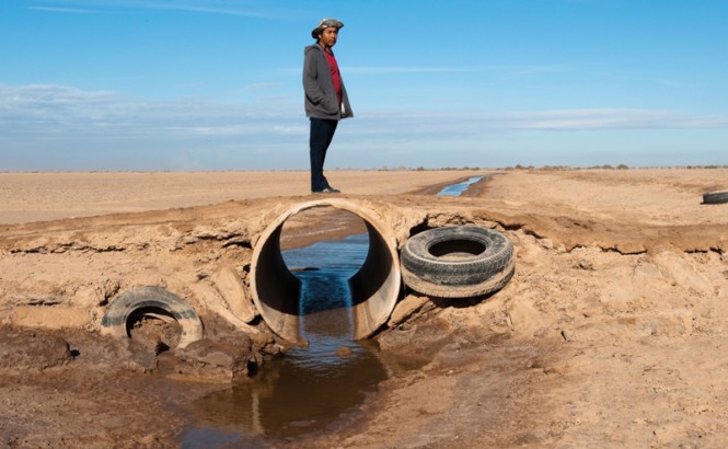 Against a blue sky with thin clouds, a male member of the Cocopah Tribe, wearing a hat, red t-shirt, dark sweater, and black pants, stands on a dirt road that sits atop a large irrigation pipe. The rest of the scene consists of an arid, brown landscape and a small stream of water that leads to the pipe, passes through it, and exits out the other side. Old tractor or car tires are placed against the dirt at either side of the pipe, and a third is seen in the distance.