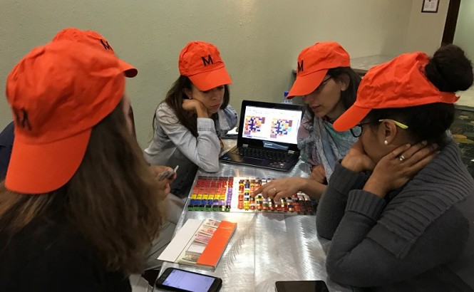 A group of participants in a Lincoln Institute course wear orange hats and gather around a game board. The hats say "M" for "middle class."