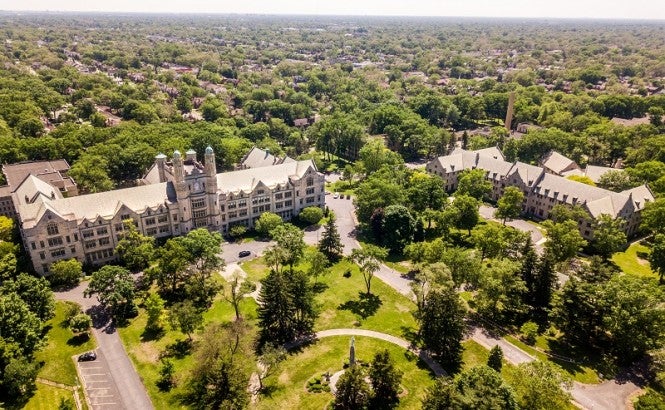 An aerial view of the Marygrove College campus and surrounding neighborhood. The campus and neighborhood have many trees. The two most visible buildings on the campus are old, large, and made of light stone with many lead paned windows. One of the buildings has a clock tower.