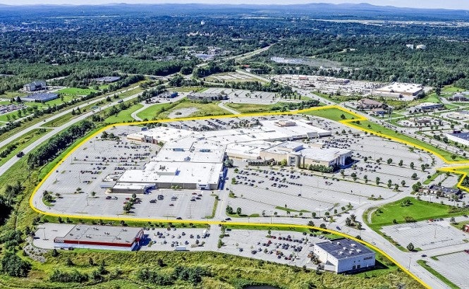 Photograph shows an aerial view of a landscape with sky and blue mountains in the distance, and a large, low, spread out building surrounded by parking spaces. Retail stores around the building and parking lot give way to suburban and rural land with many dark green trees. 