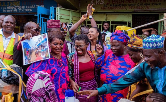 A group of people wearing brightly colored, traditional Sierra Leonean clothes laugh in a market. Freetown Mayor Yvonne Aki-Sawyerr is in the middle.