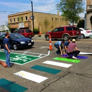 This picture shows several people laying down a new colorful crosswalk on a paved road. 