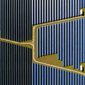 Dark blue vertical lines of solar panels seen from directly above fill a little more than a third of the left side of the image, while middle blue vertical lines of solar panels fill the remainder. A dirt road with green between it and the panels crosses the image from the left and branches into two roads in the middle and cross at angles to the top right and bottom right.