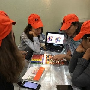 A group of participants in a Lincoln Institute course wear orange hats and gather around a game board. The hats say "M" for "middle class."