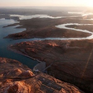 The sun glares white in the top right corner over a landscape of river and desert viewed from above. Two main branches of the Colorado river appear silver as they wind across the land and reflect the sun. They merge with the irregular, dark blue lake formed by a dam near the bottom center of the image. Roads also cross the landscape and run over the river near the dam. The terrain is a reddish brown, darker where it is in shadow.