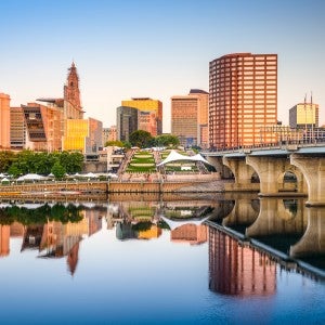 An urban skyline with tall buildings in the background, a bridge on the right and a river below, in which a reflection of the skyline is visible.