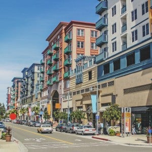 A street sits in the foreground with buildings of about 5-10 stories behind it. there are cars on the street and a few people on the sidewalks.