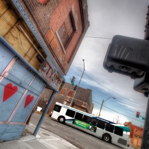 Photograph shows the side of a blighted building on a city street with crumbling paint, graffiti, and a broken store sign. A new bus drives down the street nearby. Credit: Eric Bowers.