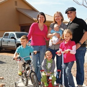 A family consisting of an older woman, older man, woman in her middle years, and four young children stands in front of a beige one-story home with a peaked roof. The older woman holds a baby and the other children are on bicycles and a scooter. There is a truck in the gravel driveway behind them.