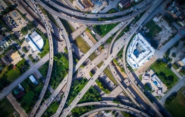 An aerial view shows a highway diagonally across the image, with on and off ramps forming a diamond shape over a semi-urban landscape of small green parks, parking lots, and commercial real estate.