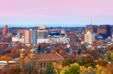 Trees with green, yellow, and red leaves stand in the foreground, buildings of varying sizes--up to 20 or 30 stories--are in the main part of the frame, and rolling hills are visible in the background.