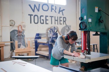 Female wood worker at a table saw in a workshop with a banner that reads working together in the background. 