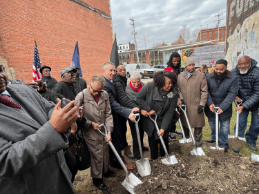Groundbreaking ceremony A group of people with shovels in a groundbreaking ceremony between two buildings with state and United States flags in the background.