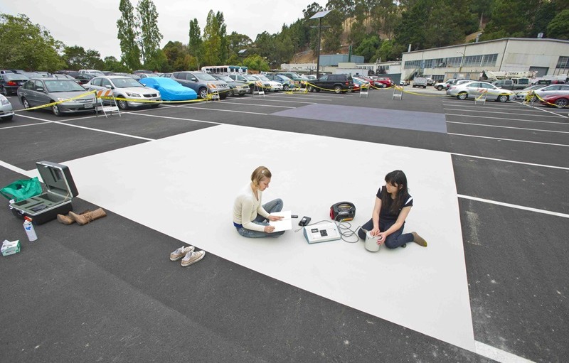 Two people sit on a square of white paint in a paved parking lot