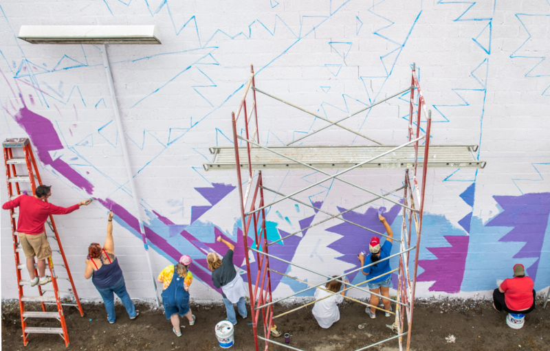A group of people painting a blue and purple mural on a white wall. A group of people painting a blue and purple mural on a white wall.