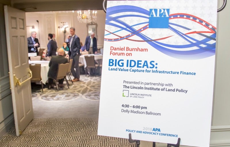 Credit: Pixelme Studio A placard occupies the right side of the frame. It reads Daniel Burnham Forum on Big Ideas: Land Value Capture for Infrastructure Finance. On the left side, people are milling around inside a room, viewed from a distance through a doorway.
