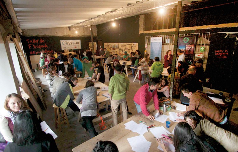 Photo shows the interior of a open room -- C-Innova's makerspace in Bogotá -- with several work tables and at least 30 people standing, sitting, and leaning on the tables as they collaborate on projects. 