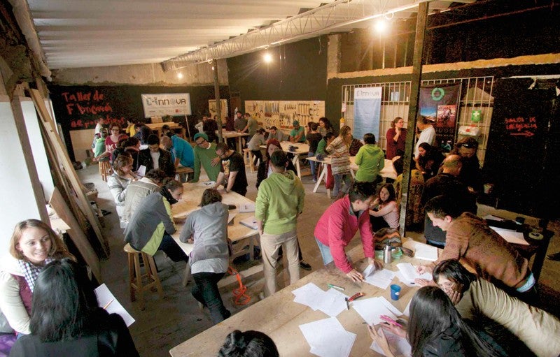C-Innova’s makerspace in Bogotá hosts workshops, classes, and meetings. Credit: C-Innova Team Photo shows the interior of a open room -- C-Innova's makerspace in Bogotá -- with several work tables and at least 30 people standing, sitting, and leaning on the tables as they collaborate on projects.