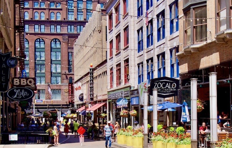 People walk on a pedestrian-only street with shops and restaurants on the sides, and tall buildings in the background.