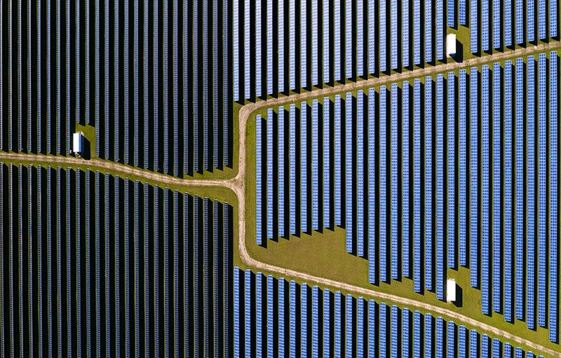 Dark blue vertical lines of solar panels seen from directly above fill a little more than a third of the left side of the image, while middle blue vertical lines of solar panels fill the remainder. A dirt road with green between it and the panels crosses the image from the left and branches into two roads in the middle and cross at angles to the top right and bottom right.