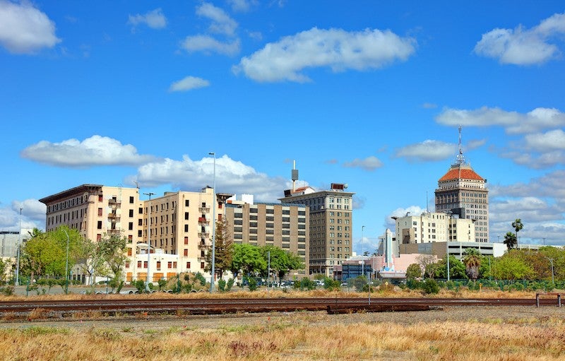 Dry grass in the foreground, train tracks mid-frame, and mid-rise buildings in the background with a blue sky behind them.