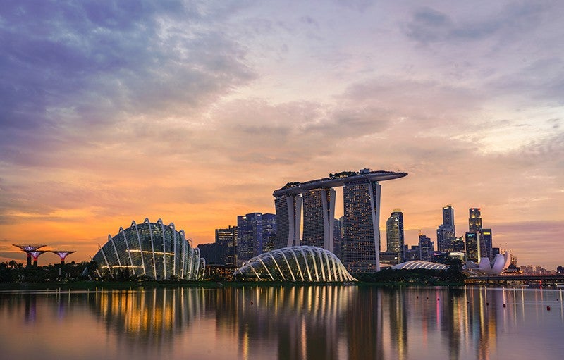 Photograph shows a dramatic urban cityscape along a body of water. The buildings are reflected in the water and the sun is setting, so the sky and clouds are orange, blue, and purple.