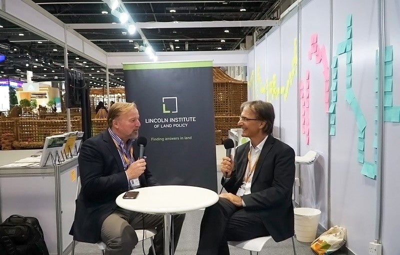 Anthony Flint and Claudio Acioly at World Urban Forum 10 Two men wearing suit jackets, Anthony Flint and Claudio Acioly, sit facing each other at a small white table and hold microphones. They are talking and smiling.