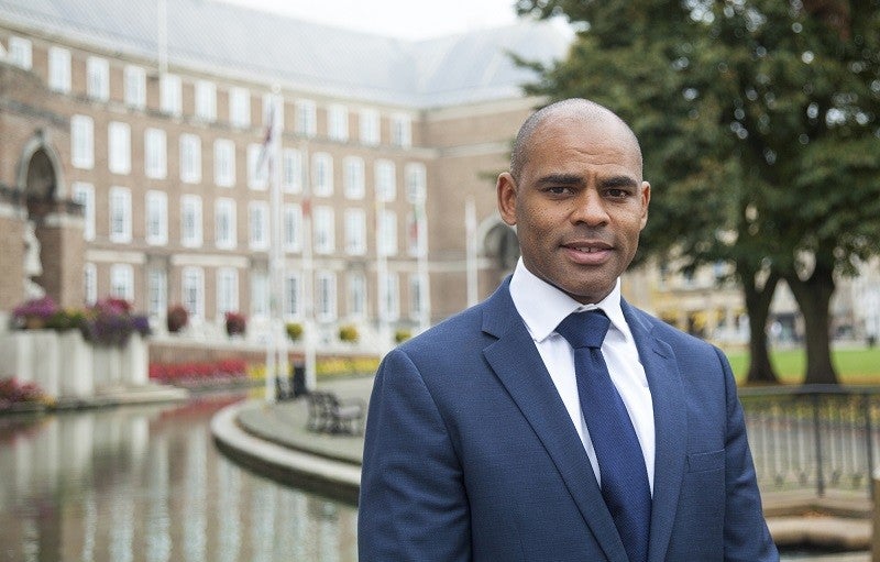 Marvin Rees. Crédito: Oficina del alcalde de Bristol. La fotografía muestra la cabeza y el torso superior de un hombre vestido con un traje azul con camisa blanca y corbata azul. Se para afuera frente a un edificio municipal con una fuente y un árbol frente a él.