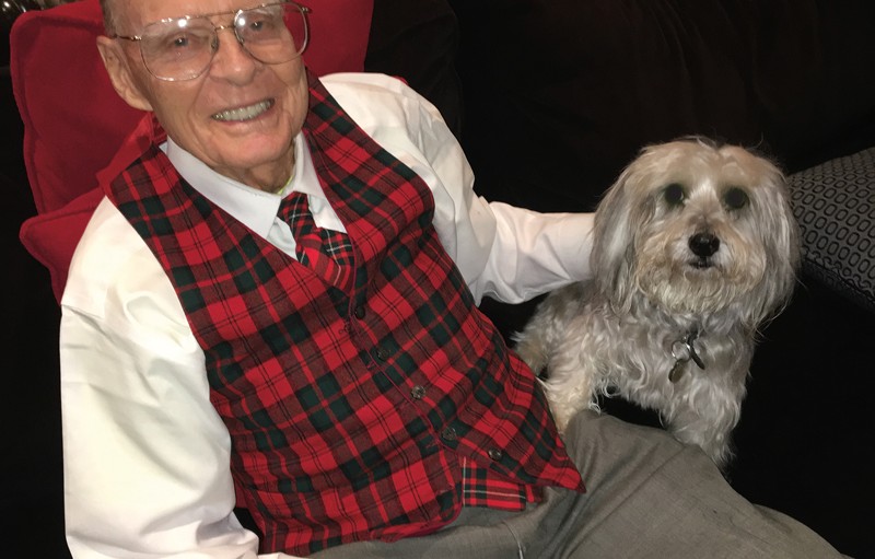 Photograph of David C. Lincoln wearing a red and black checkered vest and a white shirt, sitting on a couch in his home, with his hand resting on his small white dog, Pepper