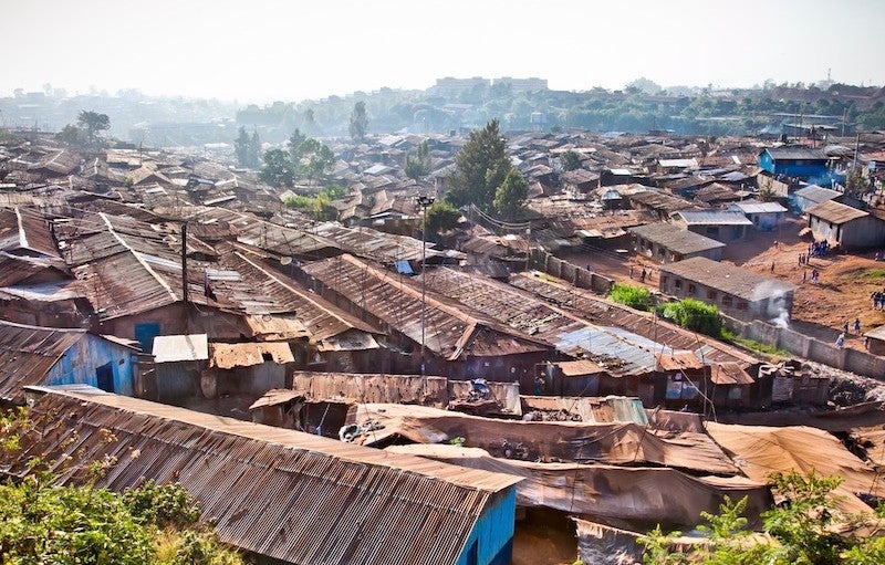 Shacks with corregated metal roofs are crowded together in most of the foreground, with a hazy urban skyline visible in the background.