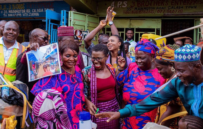 A group of people wearing brightly colored, traditional Sierra Leonean clothes laugh in a market. Freetown Mayor Yvonne Aki-Sawyerr is in the middle.