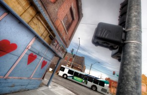 Photograph shows the side of a blighted building on a city street with crumbling paint, graffiti, and a broken store sign. A new bus drives down the street nearby. Credit: Eric Bowers.