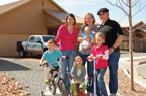 A family consisting of an older woman, older man, woman in her middle years, and four young children stands in front of a beige one-story home with a peaked roof. The older woman holds a baby and the other children are on bicycles and a scooter. There is a truck in the gravel driveway behind them.