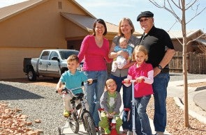 Una familia se reúne delante de su vivienda inclusiva en el desarrollo Old Las Vegas Highway, en Santa Fe, Nuevo México. Crédito: John Baker Photography. Una familia compuesta por una mujer mayor, un hombre mayor, una mujer de mediana edad y cuatro niños pequeños se para frente a una casa de un piso de color beige con un techo de pico. La mujer mayor sostiene a un bebé y los otros niños van en bicicleta y en scooter. Hay un camión en el camino de grava detrás de ellos.