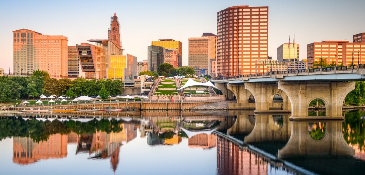 An urban skyline with tall buildings in the background, a bridge on the right and a river below, in which a reflection of the skyline is visible.