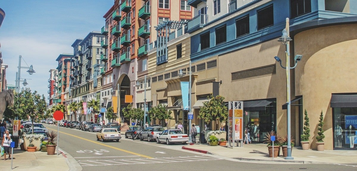 A street sits in the foreground with buildings of about 5-10 stories behind it. there are cars on the street and a few people on the sidewalks.